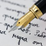 Ruka píšící klasickým plnicím perem s kovovým hrotem do linkovaného sešitu modrým inkoustem.