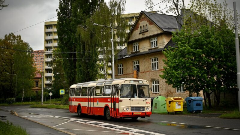 Červenobílý městský autobus Karosa ŠM 11 projíždějící ulicí se sídlištěm v pozadí