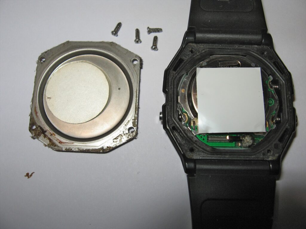 Rozebrané hodinky Casio F-91W, sejmuté kovové zadní víko, čtyři šrouby a modul v pouzdru.