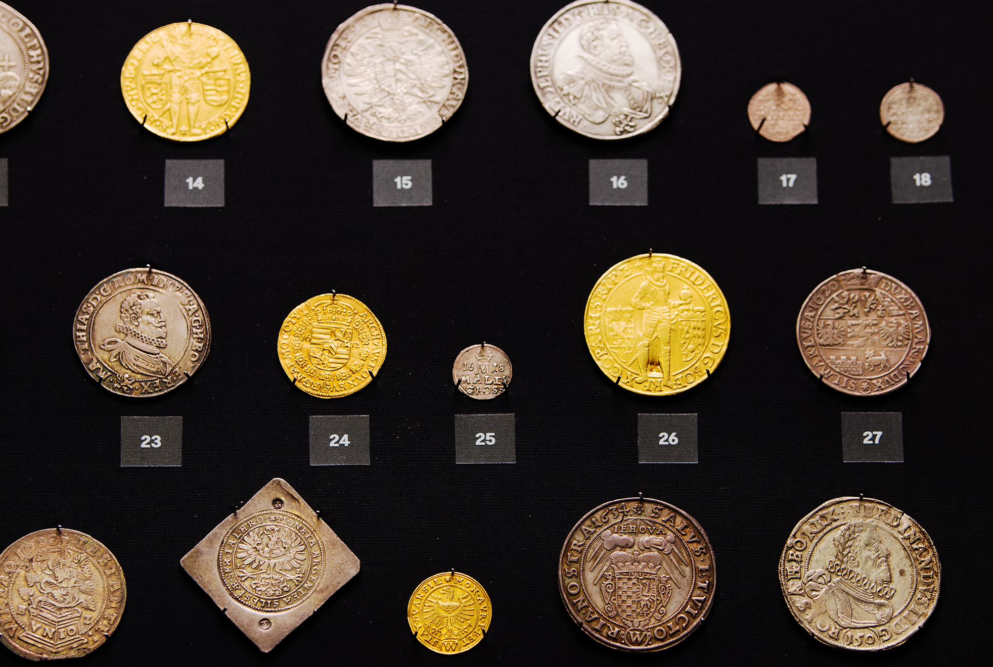 Historické zlaté a stříbrné mince vystavené na černém podkladu s čísly exponátů.
