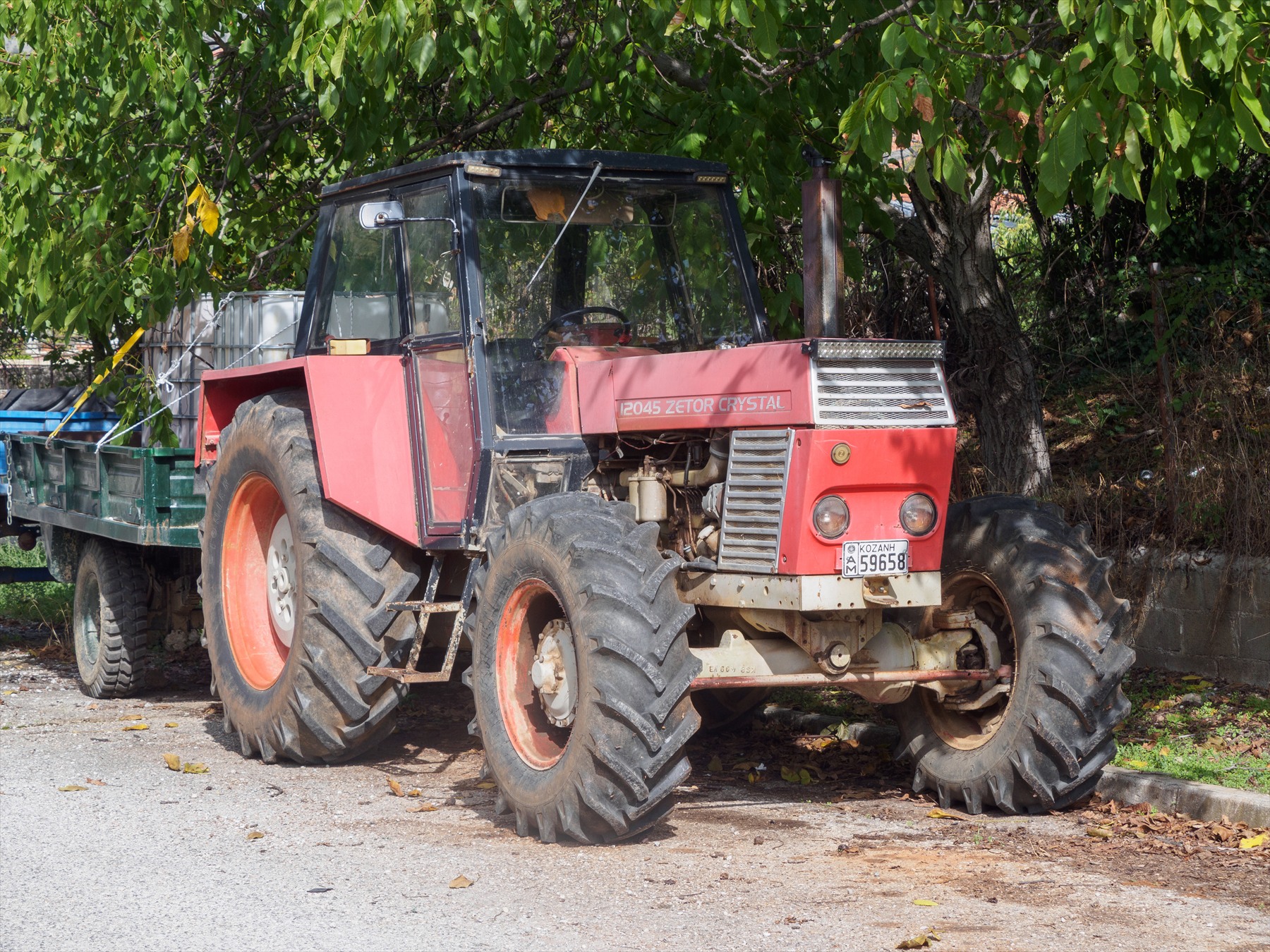 Červený traktor Zetor Crystal 12045 s pohonem všech kol a zelenou zemědělskou vlečkou na cestě se spadaným podzimním listím