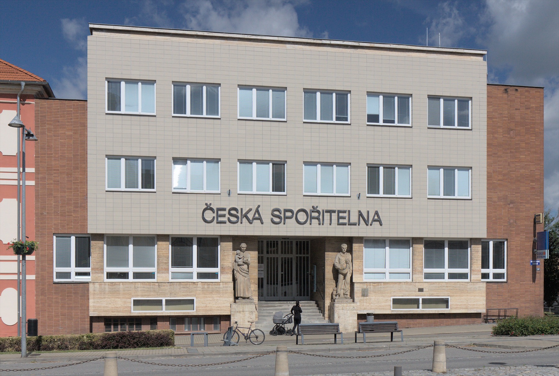 Průčelí budovy s nápisem Česká spořitelna v Napajedlech a dvěma sochami u vstupu.