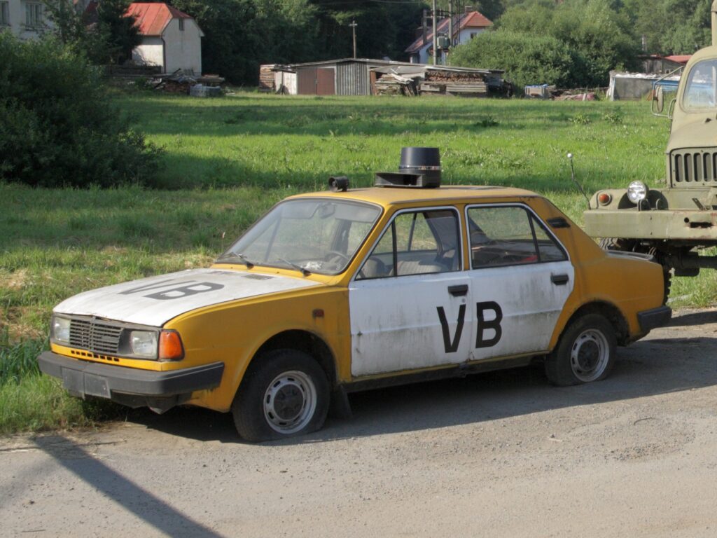 Dobový žluto-bílý automobil Škoda 105 v policejním provedení Veřejné bezpečnosti s modrým majákem na střeše.