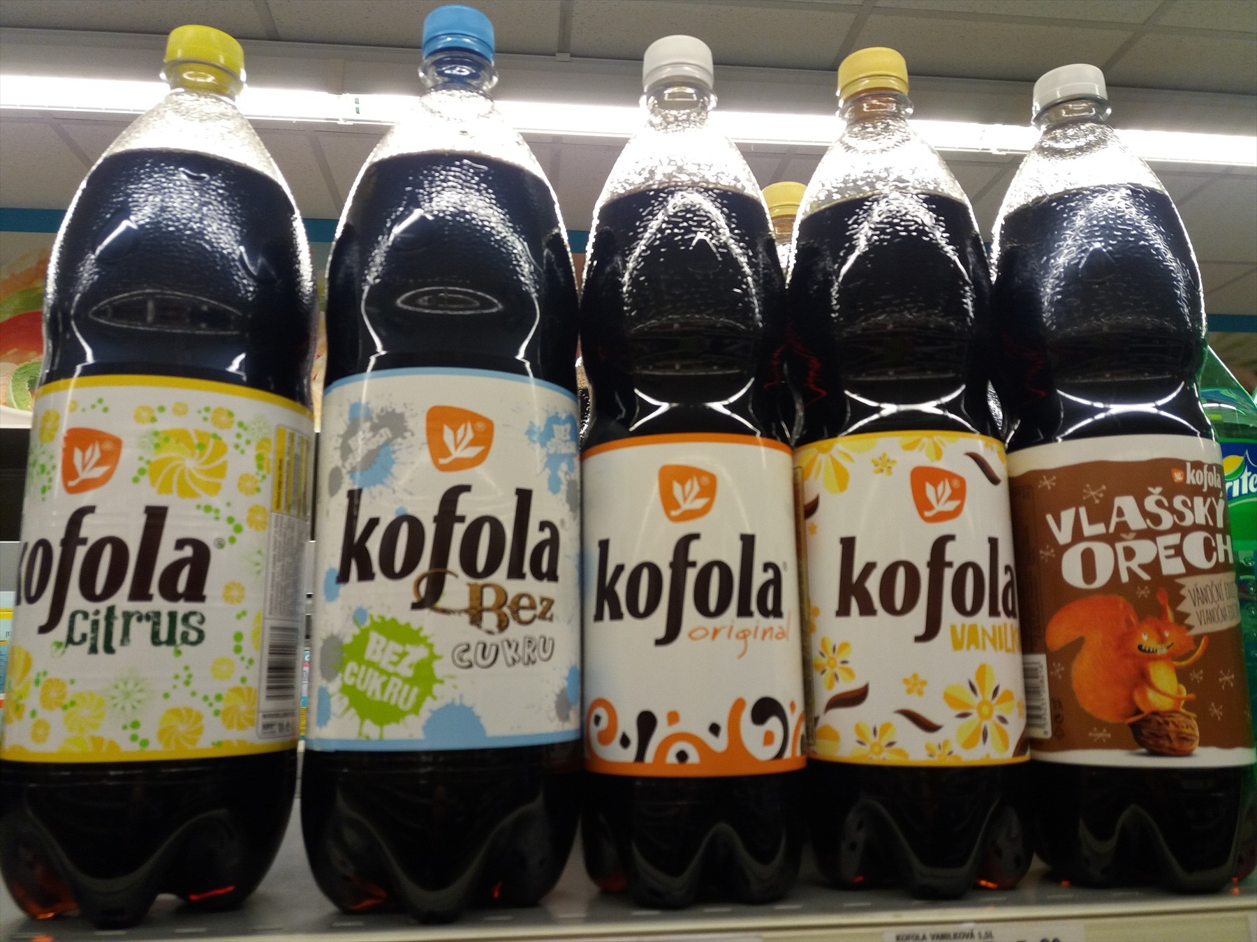 Plastové lahve Kofoly Citrus, bez cukru, Original, vanilkové a ořechové varianty na polici.