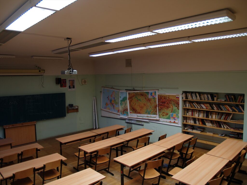 Pohled do školní učebny s řadami dřevěných lavic se židlemi, učitelskou katedrou a tabulí na stěně.