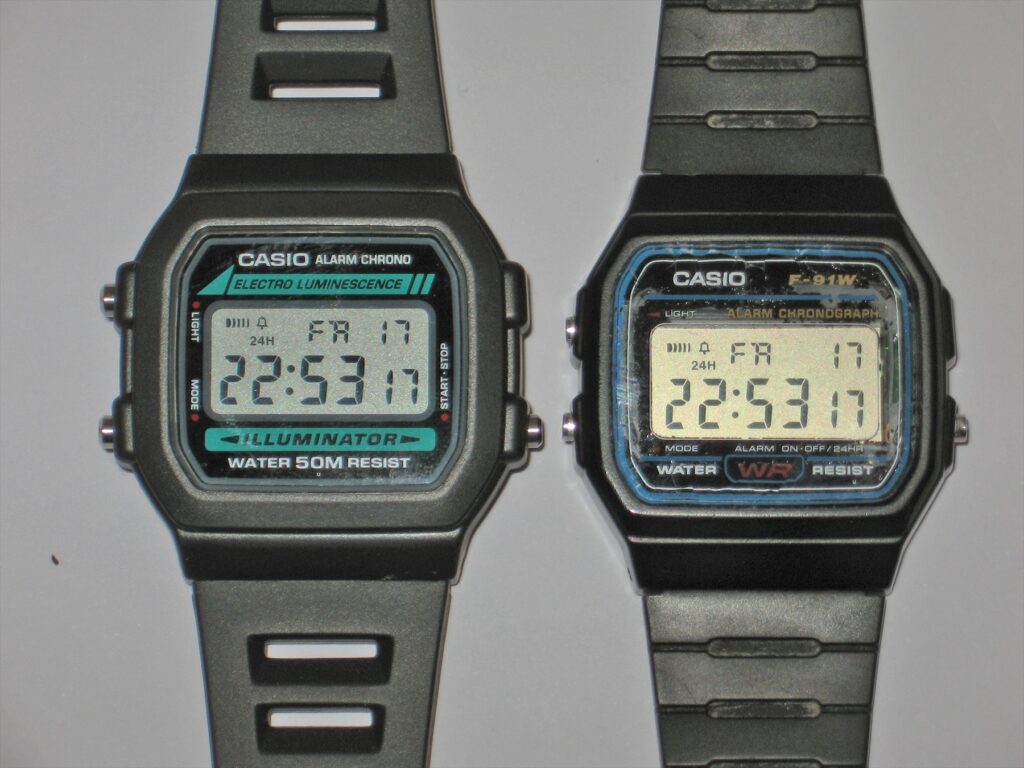 Černé digitální hodinky Casio W-86 vlevo a F-91W vpravo, položené vedle sebe.