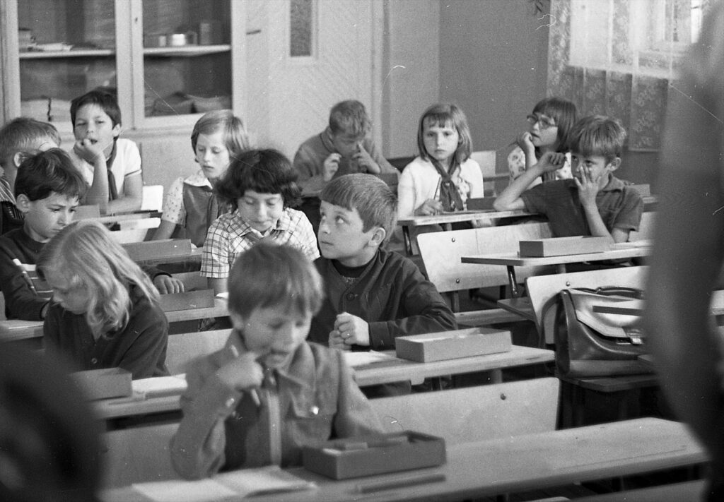 Pohled do školní třídy, kde žáci sedí v dřevěných lavicích a učitel stojí před tabulí.