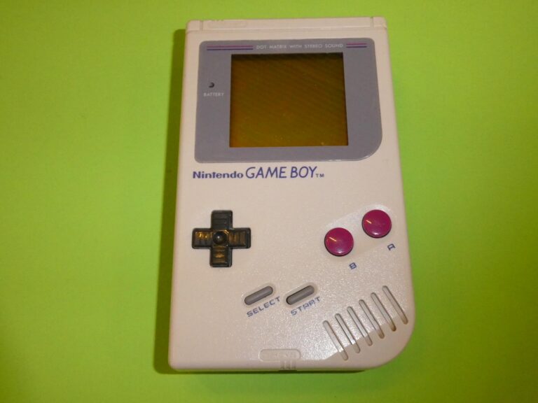 Šedý Nintendo Game Boy s tmavým displejem a fialovými tlačítky na zeleném podkladu.