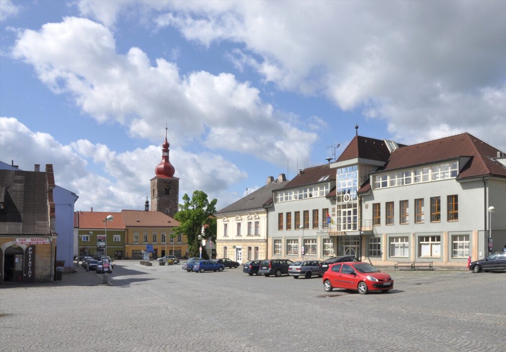 Bechyňovo náměstí v Přibyslavi s radnicí, zaparkovanými auty a věží kostela v pozadí.