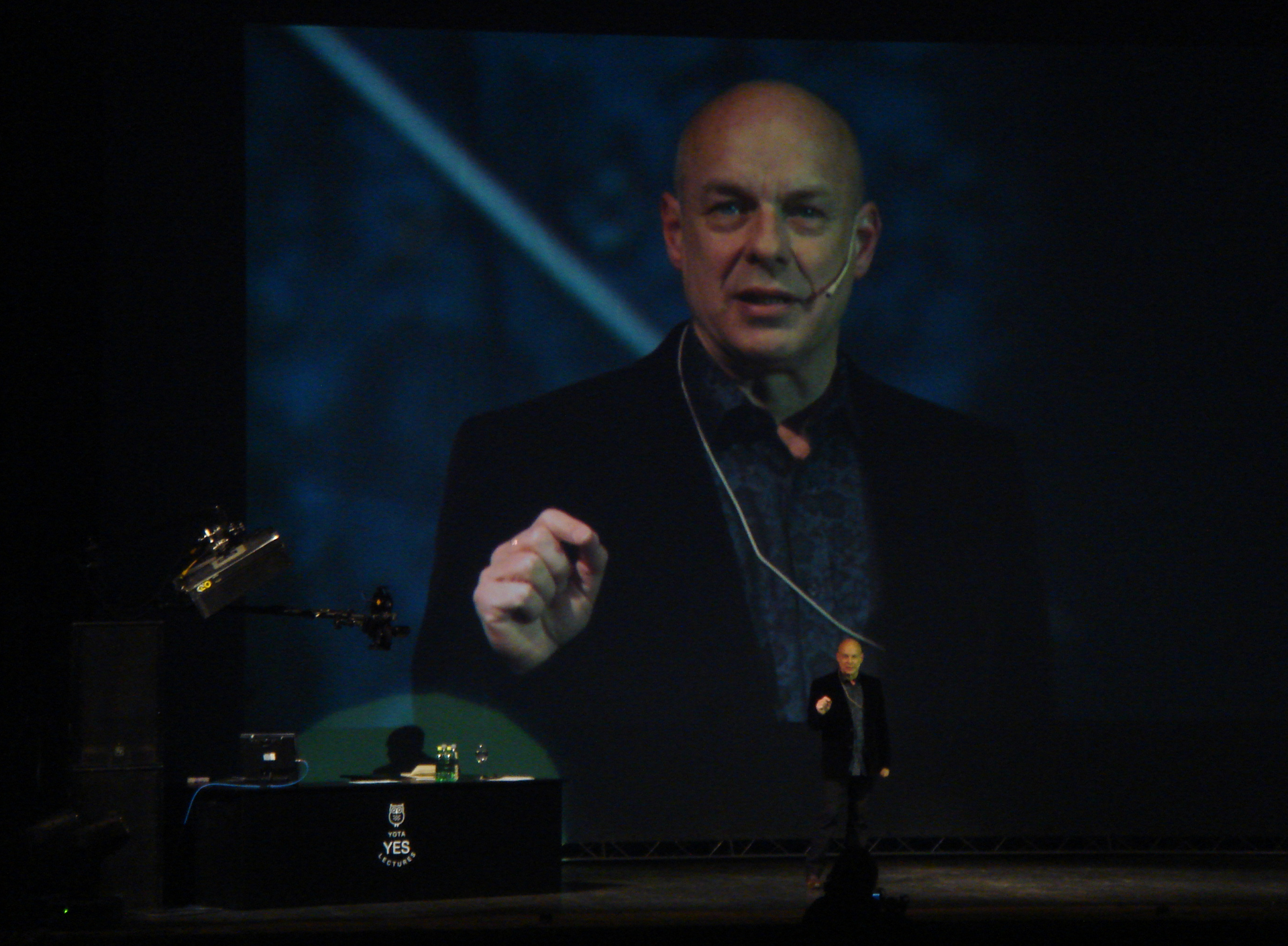 Brian Eno na pódiu před velkou projekcí svého obličeje