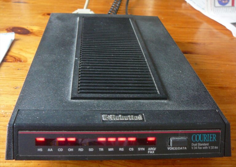 Černý modem USRobotics Courier V.34 na dřevěném stole s rozsvícenými červenými kontrolkami.