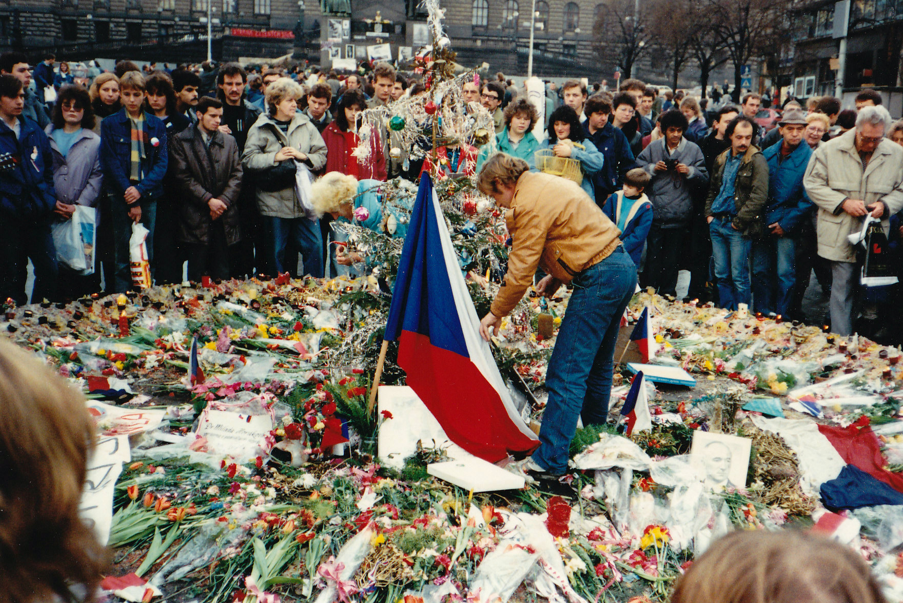 Václav Havel se sklání ke květinám a svíčkám, kolem stojí lidé s různými účesy