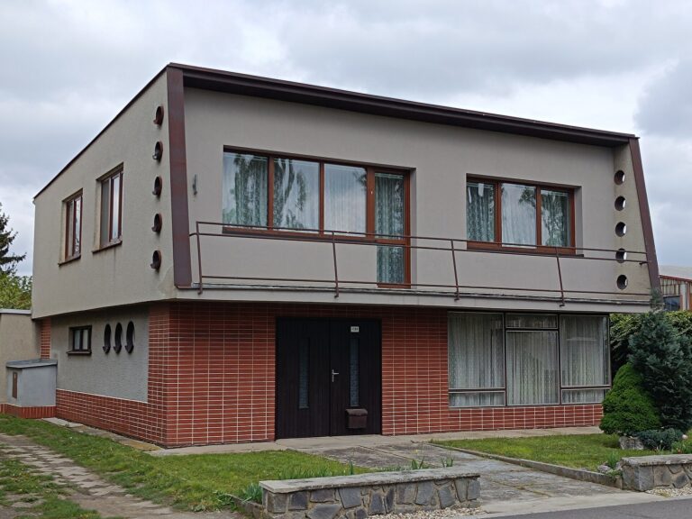 Československý patrový rodinný dům typu Šumperák s průběžným balkonem, kulatými okénky na bočnicích a cihlovým obkladem přízemí.