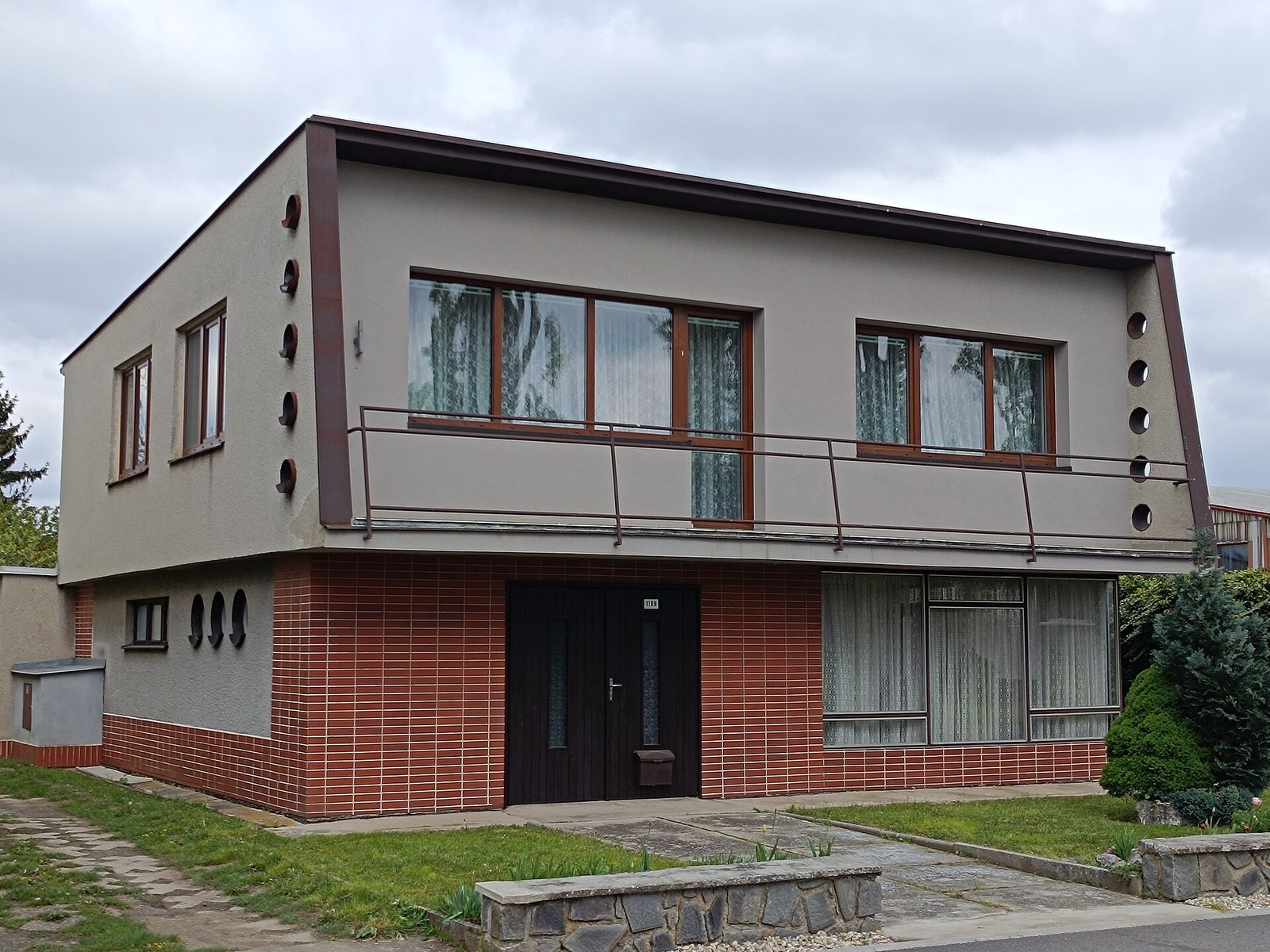 Československý patrový rodinný dům typu Šumperák s průběžným balkonem, kulatými okénky na bočnicích a cihlovým obkladem přízemí.