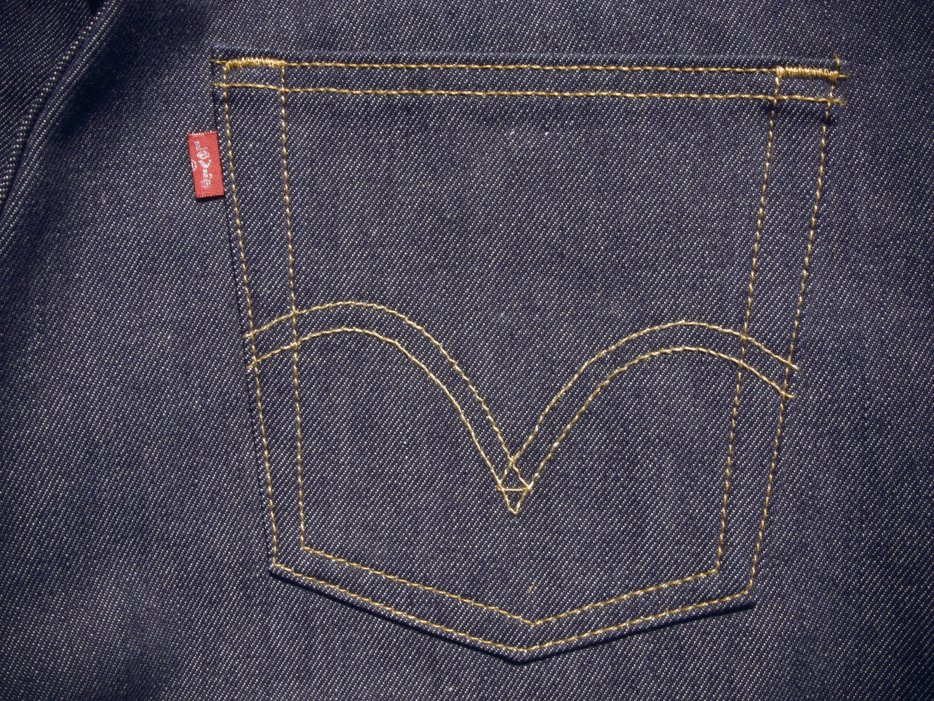 Detail zadní kapsy klasických džínových kalhot z tmavého denimu s prošíváním a červenou etiketou
