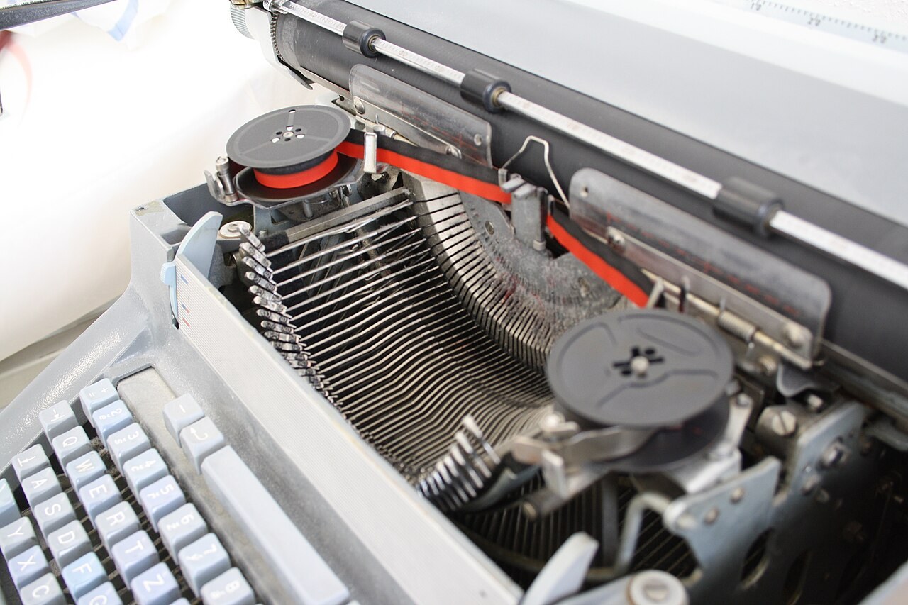 Detail typových pák s ocelovými literami a cívkového mechanismu barvicí pásky mechanického psacího stroje Consul.