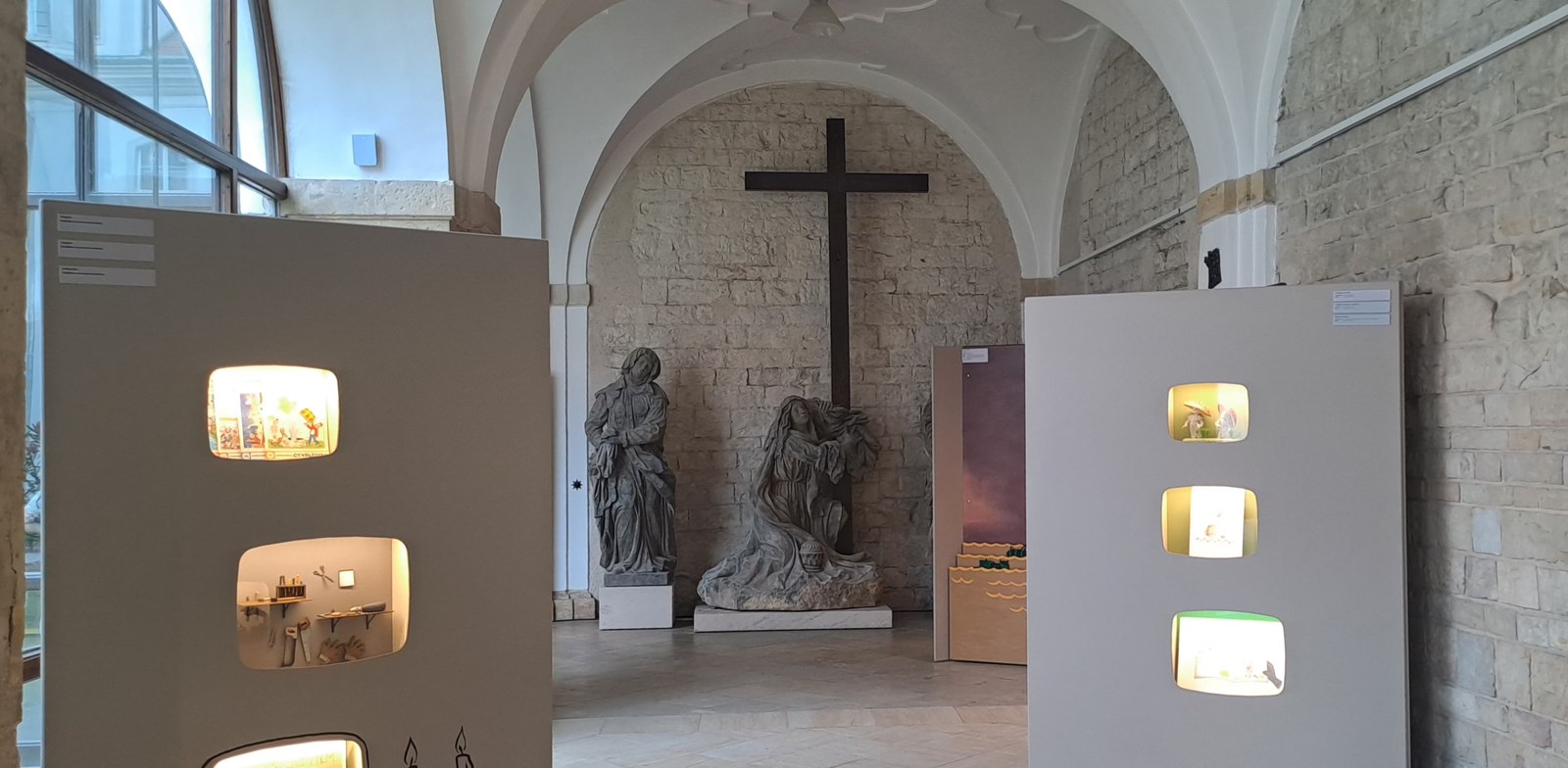 Figurka Večerníčka s papírovou skládanou čepicí sedící na autíčku s koloběžkou a koníkem v pozadí na výstavě.