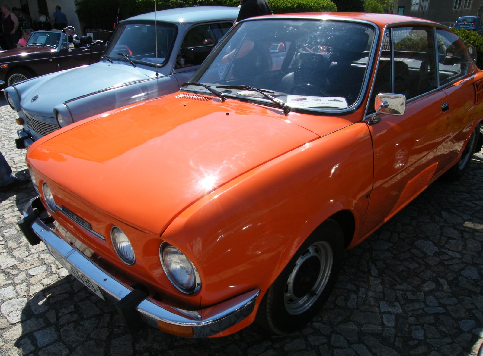 Oranžové sportovní kupé Škoda 110 R se čtveřicí kruhových světlometů a chromovaným nárazníkem zaparkované na dlážděném prostranství.