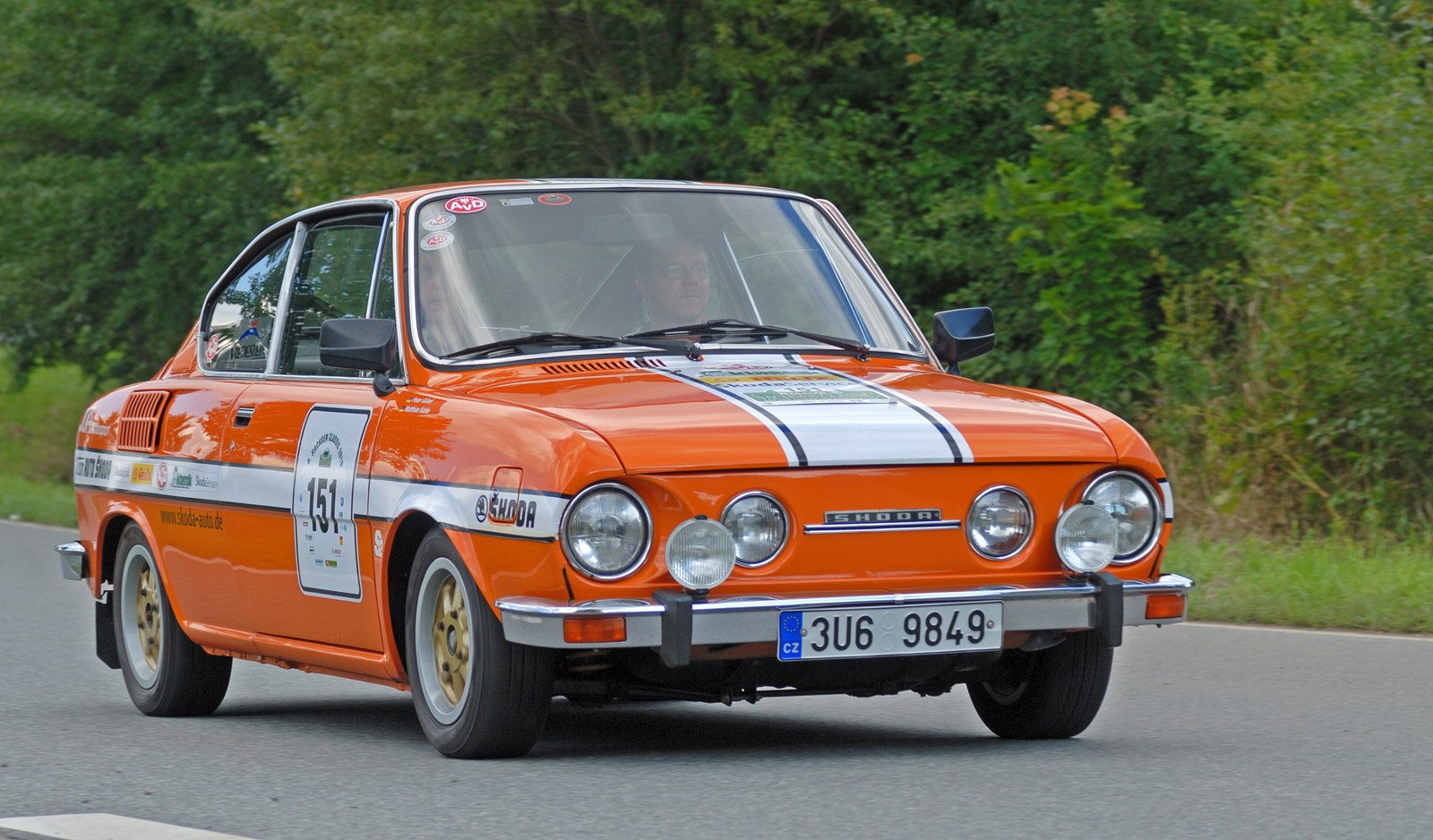 Sportovně upravená oranžová Škoda 110 R se startovním číslem a přídavnými dálkovými světly při jízdě na veteránské rallye.
